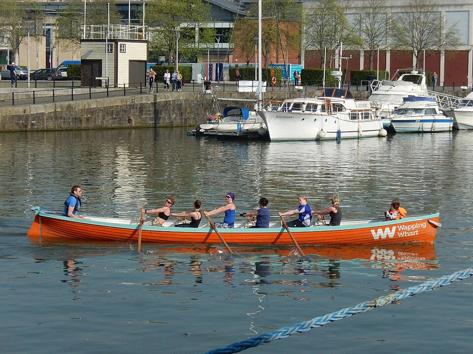Bristol Floating Harbour’da turuncu bir teknedeki kürekçiler ve kıyı; Nisan 2018.