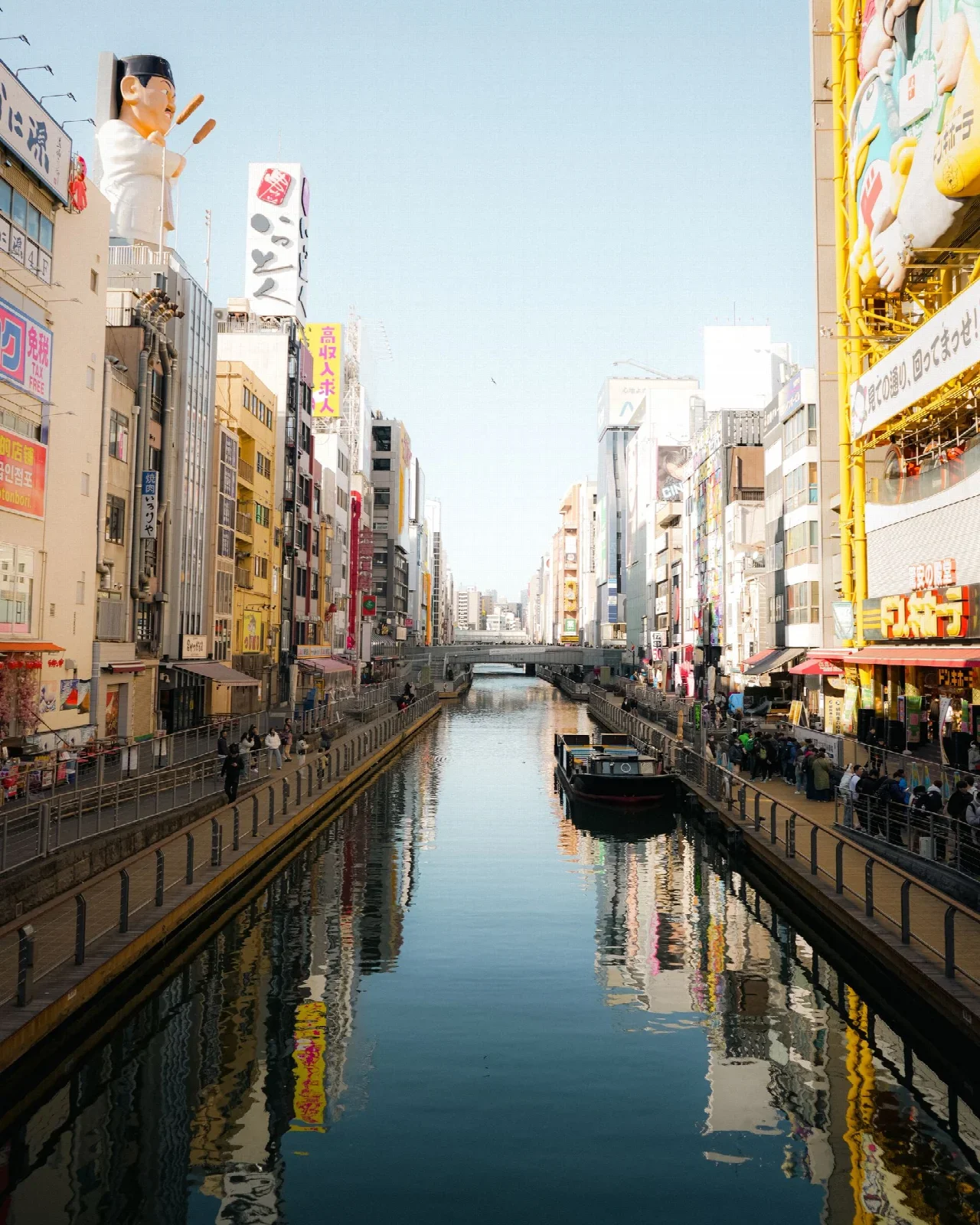 Dotonbori kanalında kıyı ve suya yansıyan binalar; Sam Tan. Öğünler arasındaki şehir yürüyüşüne bağlam.